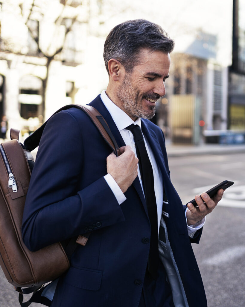 Man in business suit with a backpack walking down the street smiling at his phone.